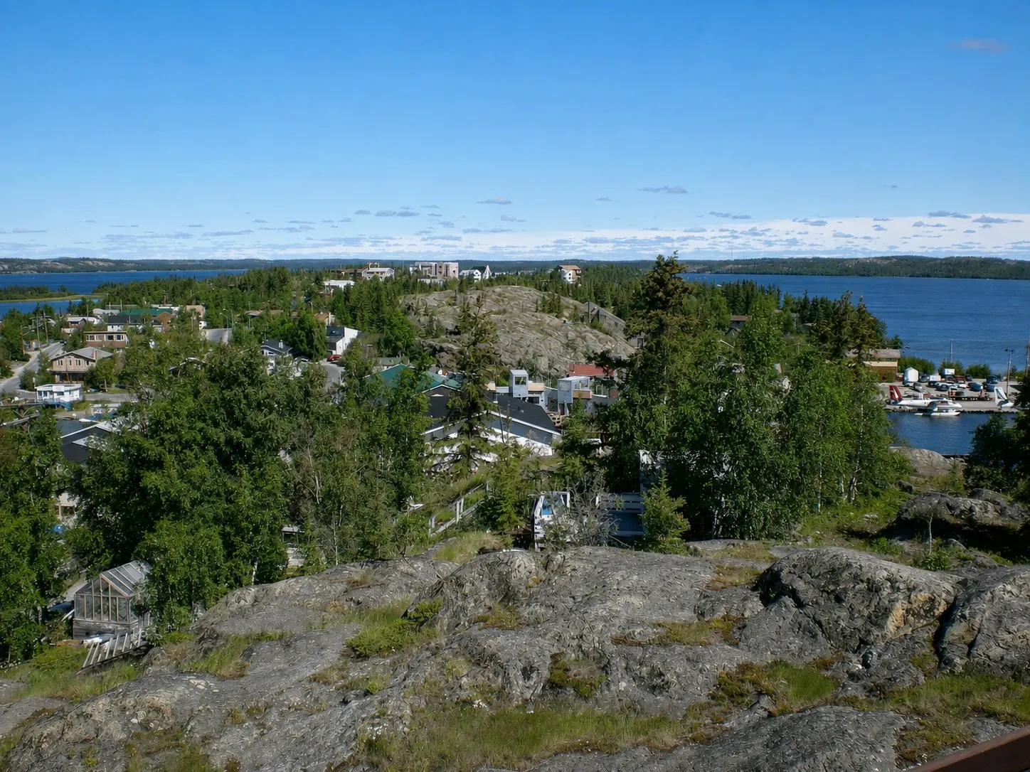 AI-enhanced view: Summer view across Yellowknife Old Town toward N’Dilo with granite outcrops, trees, houses, lake water, and floatplanes.